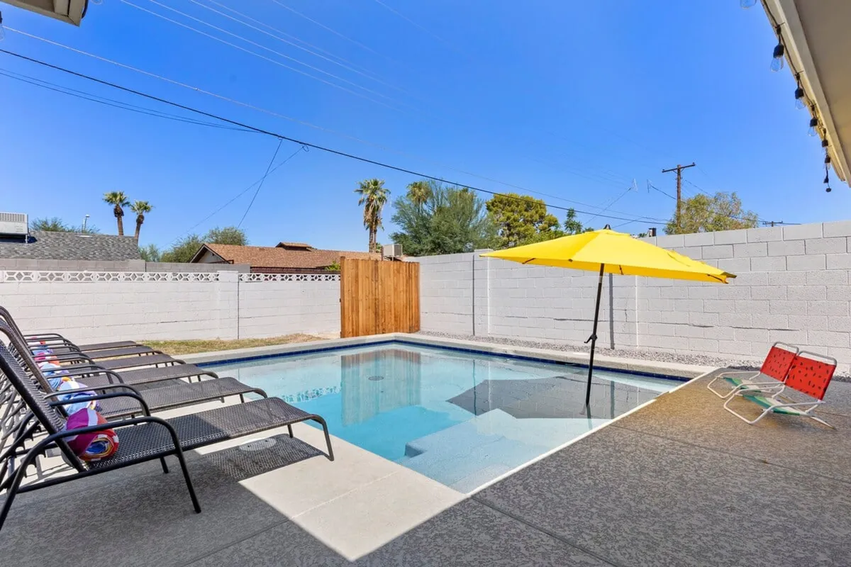 A sun-drenched backyard scene featuring a sparkling in-ground swimming pool, yellow umbrella for shade, and multiple lounge chairs arranged for relaxation. The space is open, inviting, and designed for both sunbathing and gathering, with sunset hues highlighting the tranquil, social atmosphere.
