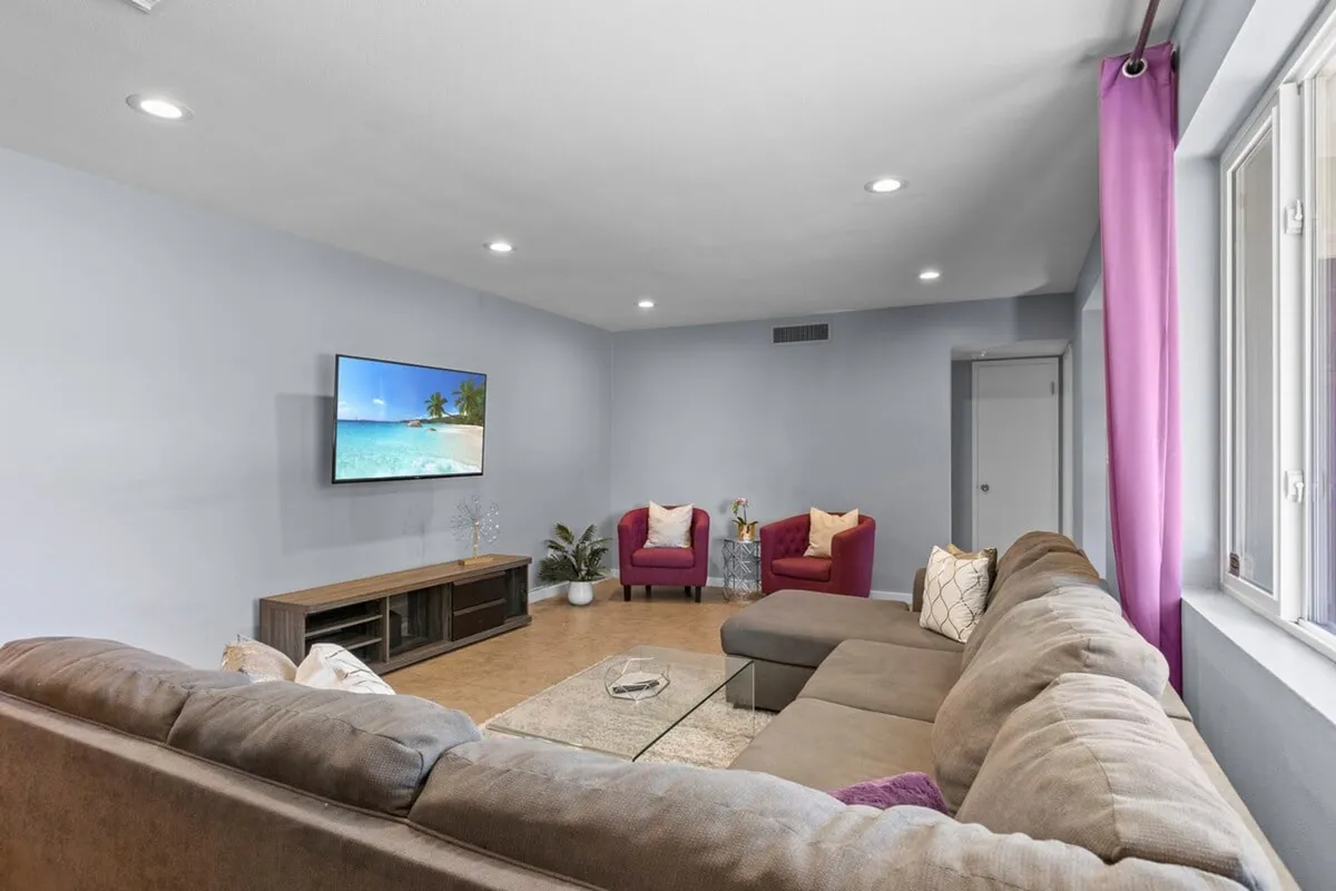 A spacious living room with a large sectional sofa positioned for socializing, a TV showcasing a tranquil beach scene, and two accent chairs. The environment is inviting and designed for relaxation, movie nights, or group conversations.