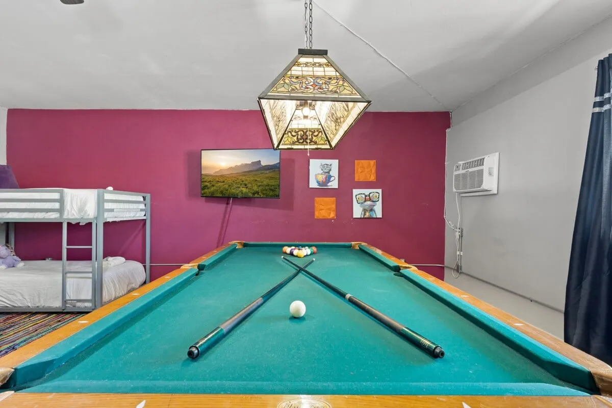 This interior view highlights a cheerful, casual room with a pool table under a stained glass light, a TV for entertainment, a bunk bed, and lively artwork. The space is designed for everything from friendly pool games to relaxed TV time, and group hangouts.