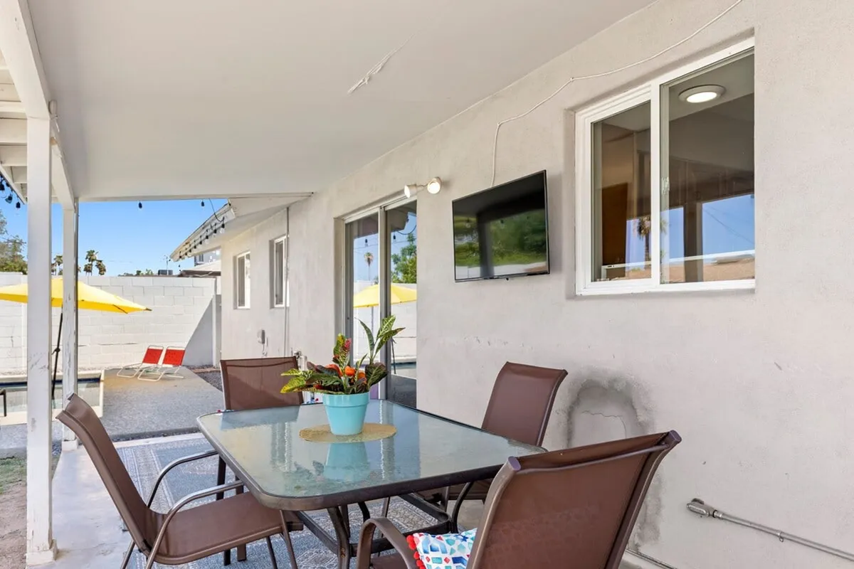 An outdoor patio setup with a dining table, potted plants, comfortable seating, and a large outdoor TV mounted nearby. The arrangement is ideal for group dining, watching the game, or casual conversation, with sunlight enhancing the welcoming atmosphere.