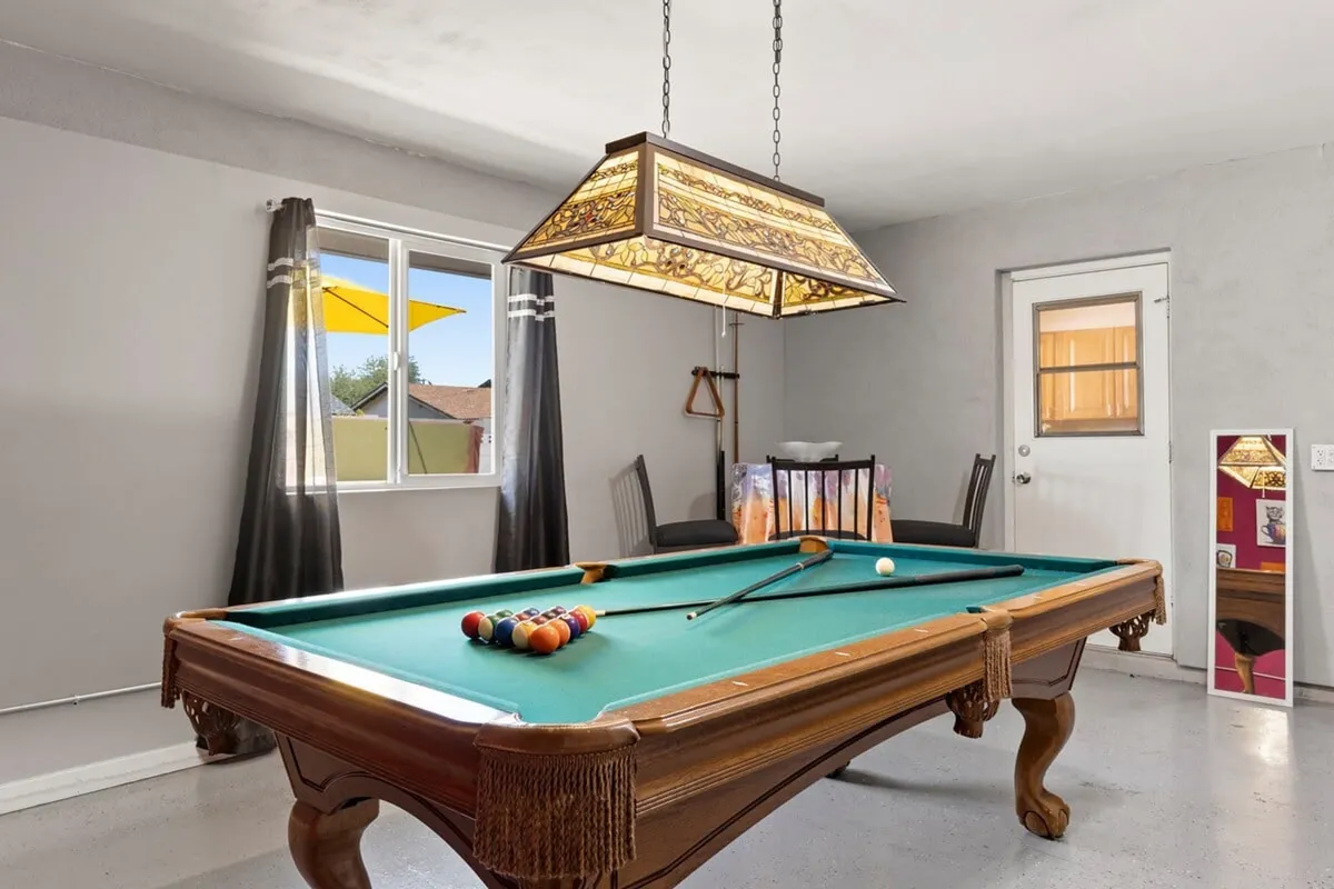 A playful game room with a pool table under a stained glass light fixture, adjacent to a bright window with a view of a yellow patio umbrella. This space invites casual socializing and friendly competition for guests looking to unwind after hours.