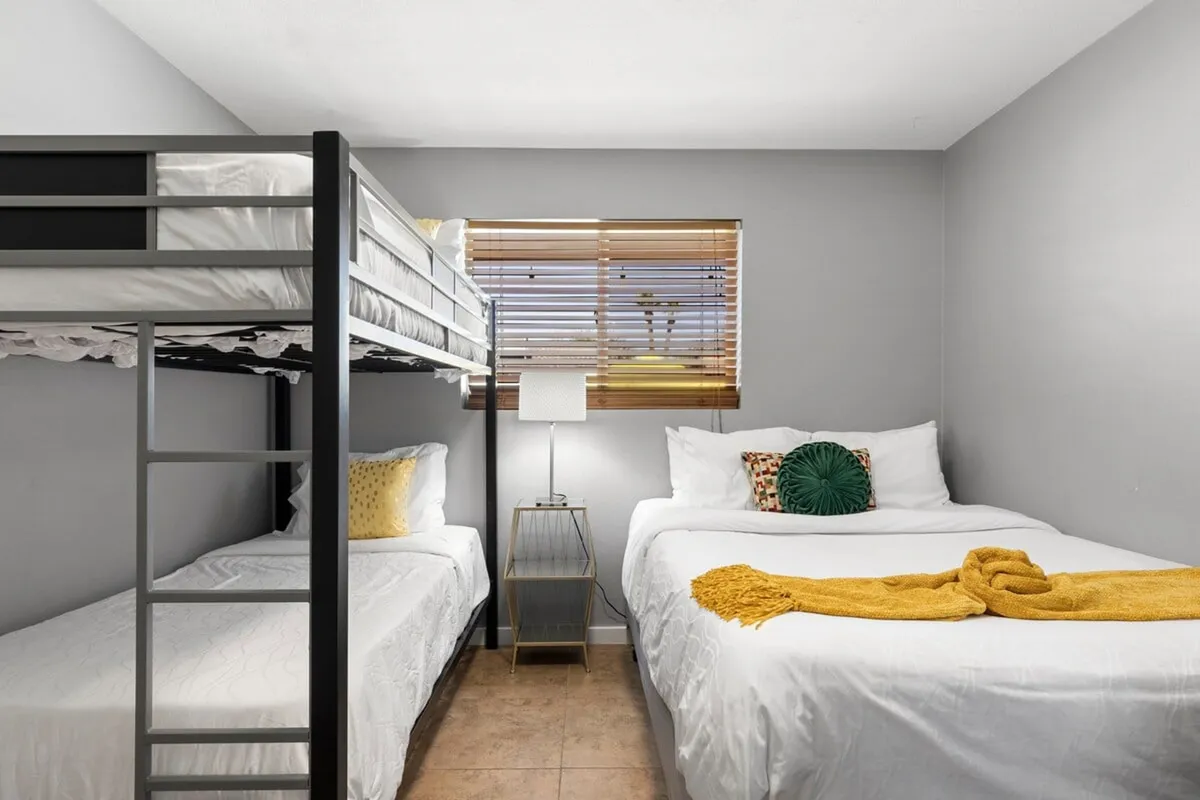 A bright bedroom featuring a bunk bed and a queen bed, adorned with decorative pillows and a lamp beside a window. The arrangement suggests comfort and flexibility for families with children or guests, and the natural light adds to the inviting atmosphere.