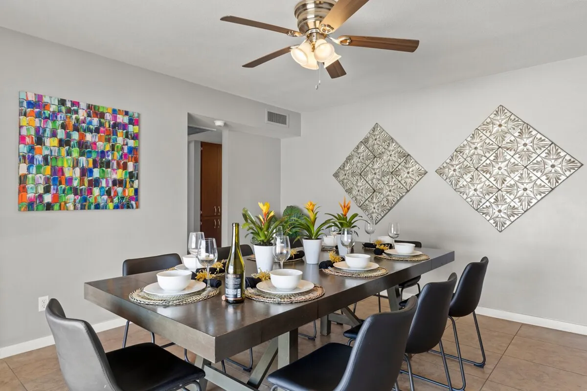 A welcoming dining area set for a lively meal, featuring a large dining table surrounded by chairs, bold colorful artwork on the wall, and playful decorative accents. The space radiates vibrancy, suggesting group gatherings, celebrations, or shared meals.