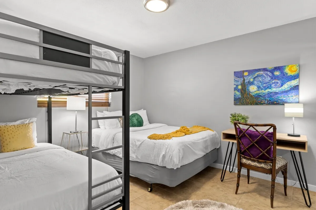 A playful and creative bedroom scene featuring a bunk bed, desk with chair, artwork on the wall, and a cheerful yellow throw blanket. The setup is ideal for children, communal stays, or work teams.