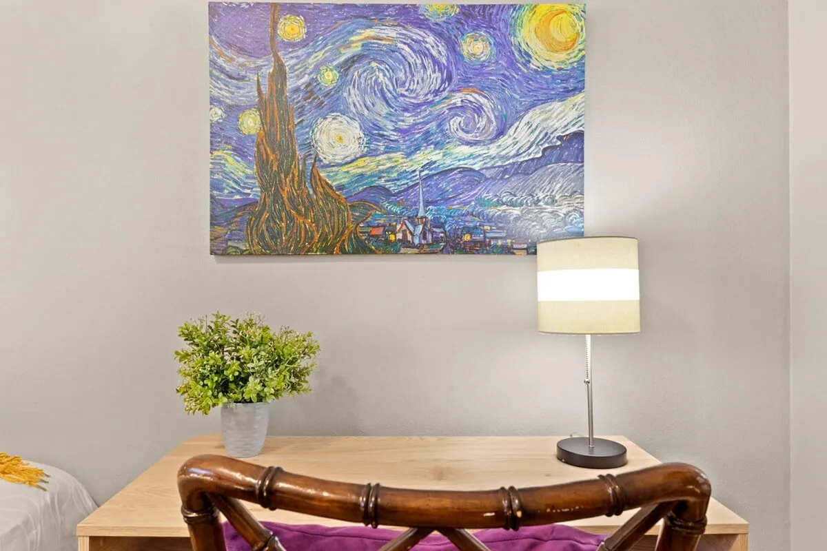 A focused, cozy workspace anchored by a desk and wooden chair under a desk lamp, with a potted plant adding freshness. A framed Starry Night painting inspires creativity, setting the mood for quiet productivity or thoughtful reflection.