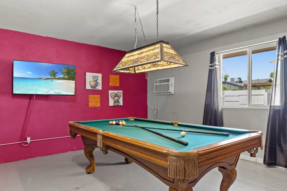 This interior image spotlights a vibrant activity zone featuring a pool table, a decorative light fixture overhead, a large TV displaying a beach scene, and colorful wall art. Natural light from the window brings the space to life and hints at its connection to the outdoors. The layout encourages both active play and relaxed movie watching.