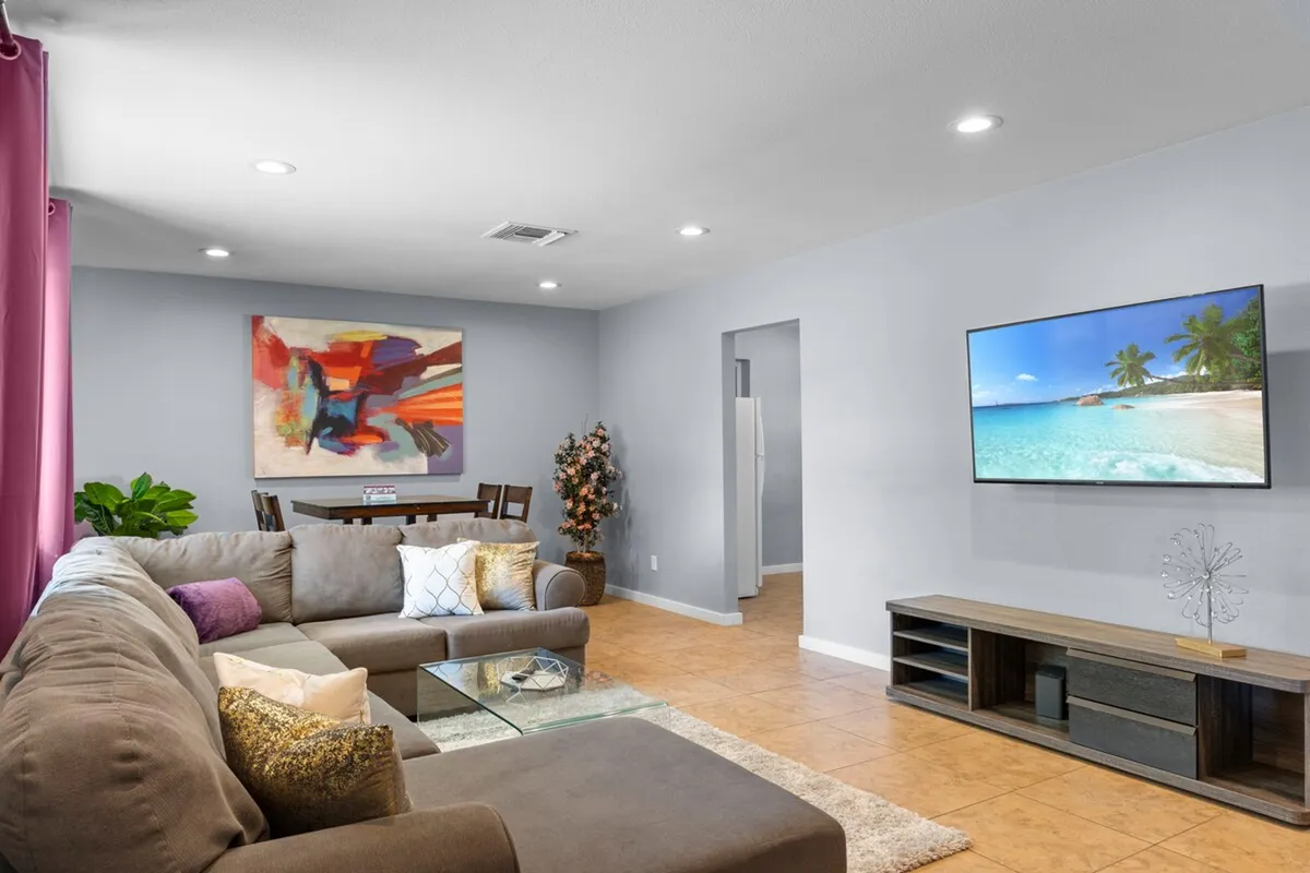 A comfortable living area featuring a large sectional sofa, abstract artwork, a flat-screen TV, and a dining table. The space is arranged for both relaxation and entertainment, encouraging movie nights or shared meals.
